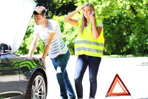 Eine Person telefoniert nach einer Fahrzeugpanne mit dem Pannendienst, während eine weitere Person das liegengebliebene Auto überprüft und die Unfallstelle abgesichert ist.